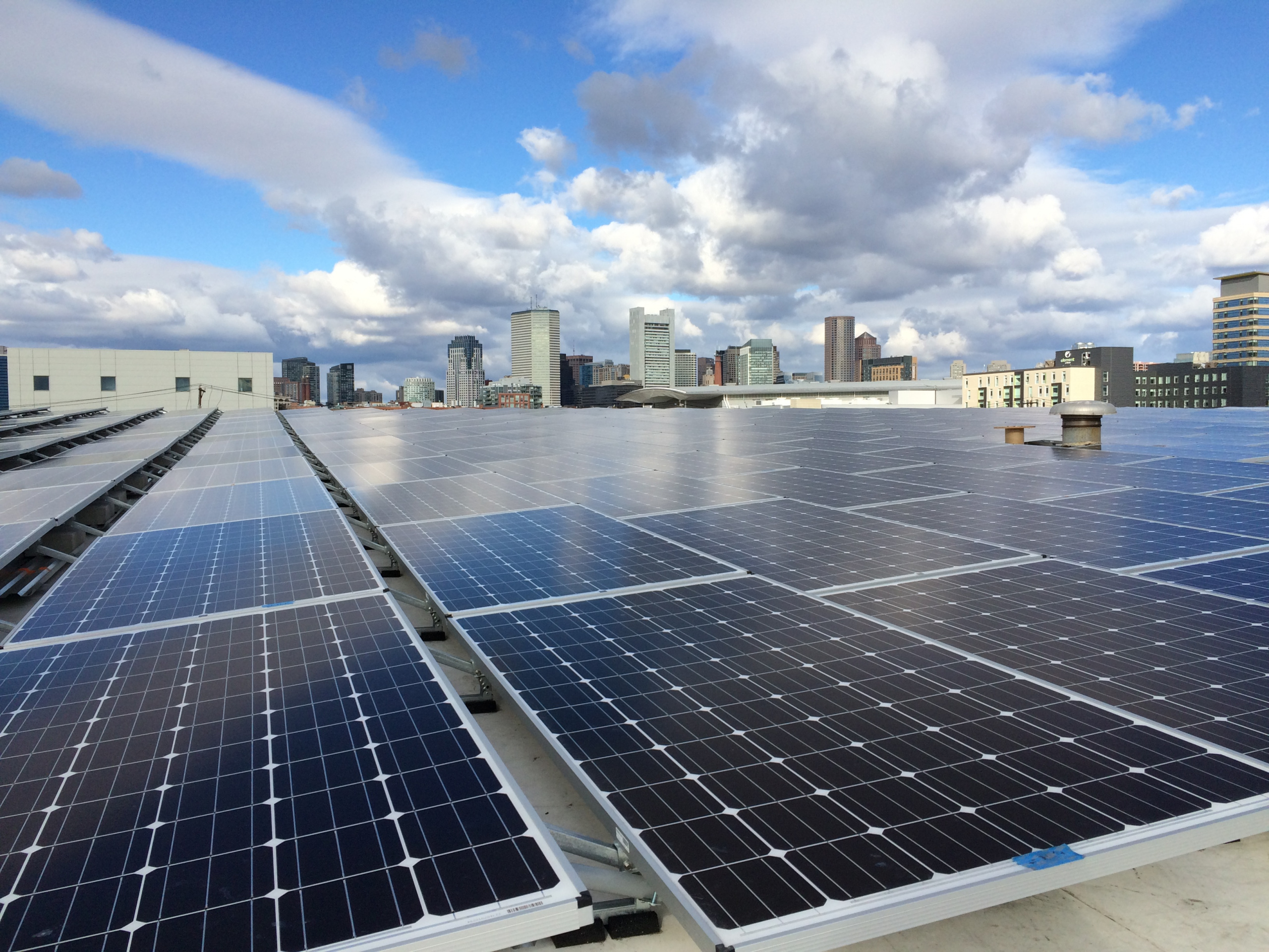 The Boston skyline is visible over the largest solar installation in Boston, located on a warehouse in the Seaport district.